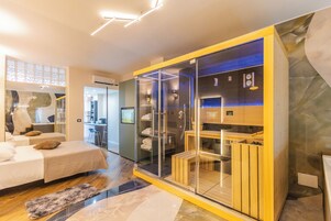 Una moderna camera d'albergo con zona doccia in vetro, una panca di legno e un angolo salotto con vista sulla cucina.