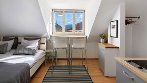 A compact room with a bed, a desk, a chair, a small kitchenette, and a window with a view of mountains. - Studio 'Sommavilla - Small' with Mountain View and Wi-Fi (Silandro)