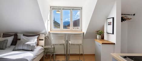 A compact room with a bed, a desk, a chair, a small kitchenette, and a window with a view of mountains.