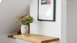 A white wall with a framed abstract painting, a wooden shelf with a vase of flowers, and a light-colored cabinet below. - Studio 'Sommavilla - Small' with Mountain View and Wi-Fi (Silandro)