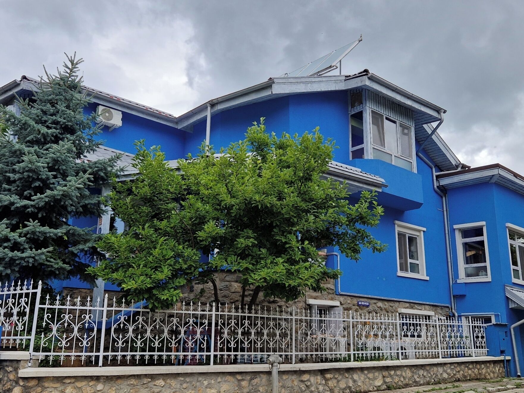 A blue building with a stone wall foundation, a metal fence, and a tree in front.