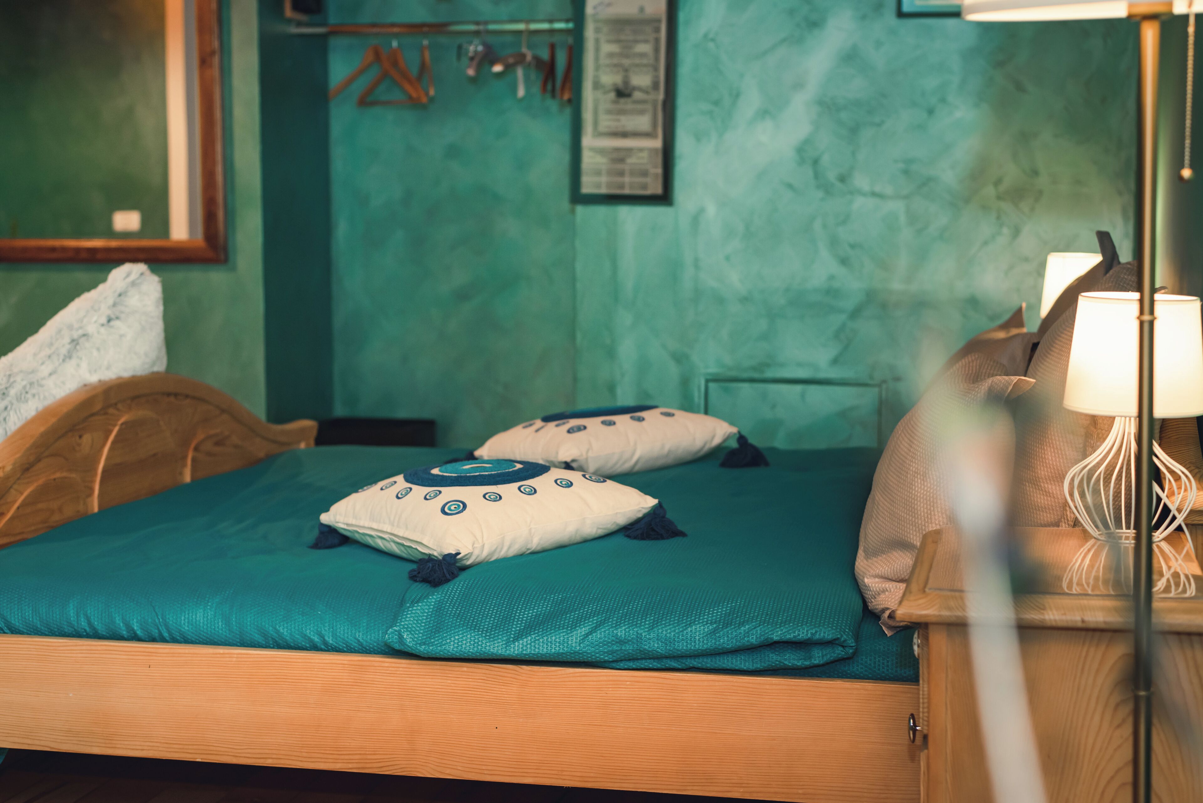 A neatly made bed with teal bedding and decorative pillows, a wooden headboard, a bedside table with a lamp, and a green wall with a framed picture and a hanging coat rack.