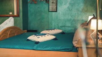 A neatly made bed with teal bedding and decorative pillows, a wooden headboard, a bedside table with a lamp, and a green wall with a framed picture and a hanging coat rack.