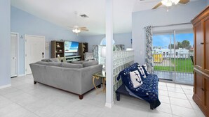 A spacious living room with a grey sofa, a TV, and a balcony with a view of a green landscape. - ☀️Steps to Beach | Shared Pool | Awesome Game Room | Beach Villa A☀️ (Fort Myers Beach)