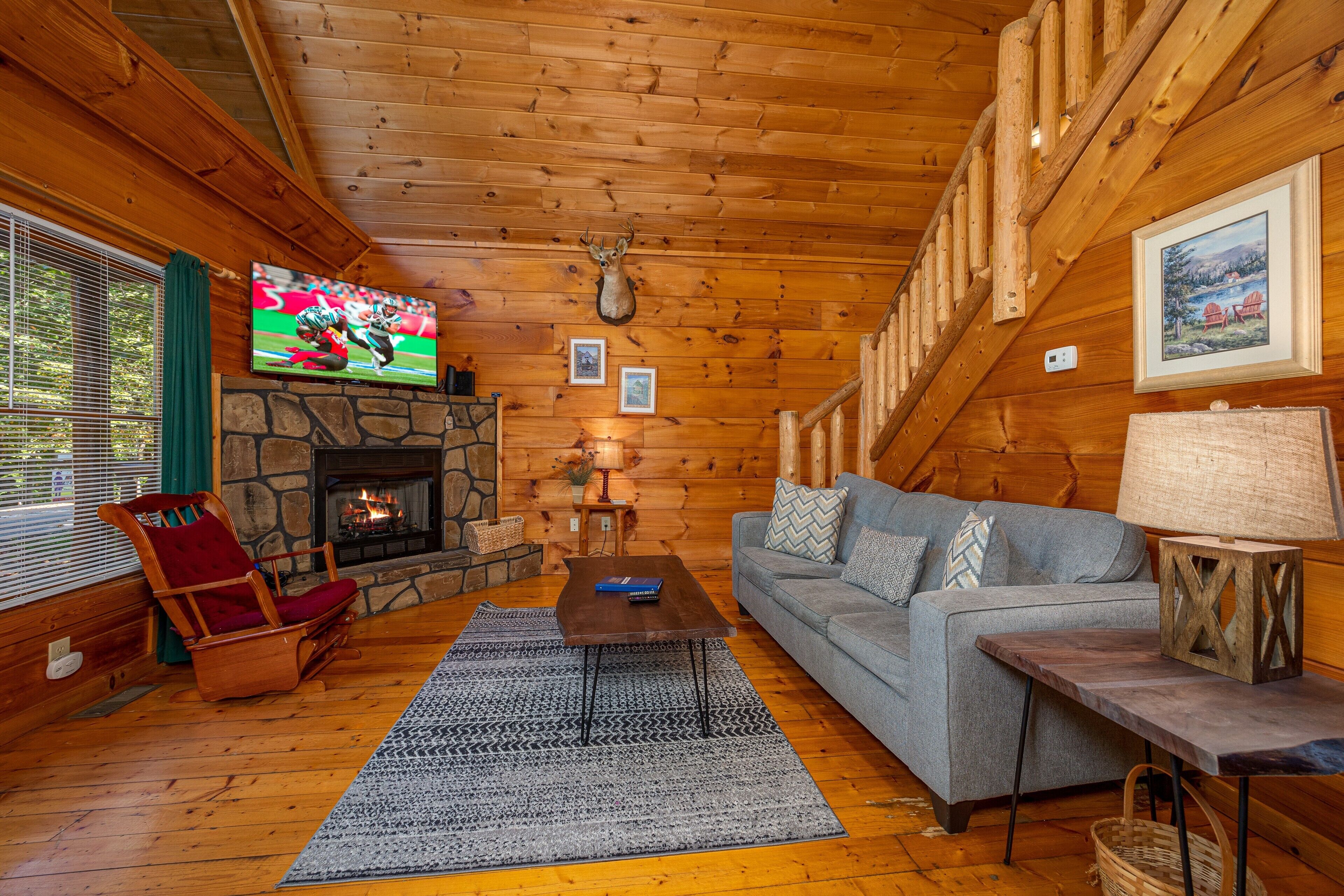 Un acogedor interior de cabaña de troncos con chimenea de piedra, un televisor montado en la pared, una cabeza de venado decorativa y una zona de estar cómoda con sofá y sillones.