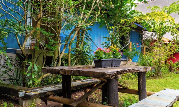 A rustic wooden table with a potted plant in the center, surrounded by lush greenery and a blue building in the background. - Pousada Eco Manguinhos (Serra)