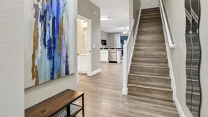 A hallway with a wooden staircase, a kitchen in the background, and a large abstract painting on the wall.