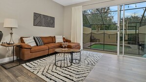 A modern living room with a brown leather sofa, two side tables, a patterned rug, and a large sliding glass door leading to an outdoor area with a pool.