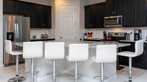 A modern kitchen with dark cabinets, stainless steel appliances, and a central island with white bar stools. - Modern 7BR Villa | Pool + Resort Amenities (Kissimmee)