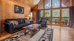 A wooden cabin interior with a leather sofa, a wooden coffee table, and a large window overlooking a deck.