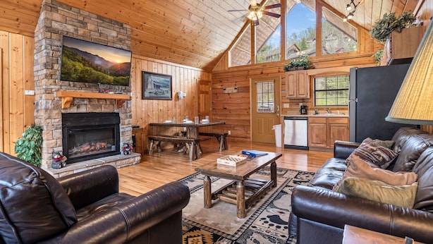 A cozy cabin interior with a stone fireplace, leather sofas, a dining table, and a kitchen area.