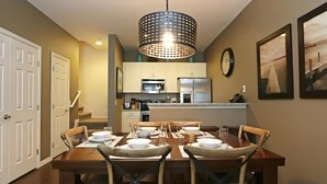 Comedor con una mesa de madera para cuatro, cocina al fondo y una lámpara colgante grande.