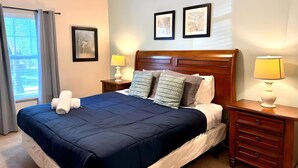 Una cama bien hecha con una colcha azul y almohadas, un cabecero de madera, dos lámparas de noche y una mesita de noche de madera con cajones.