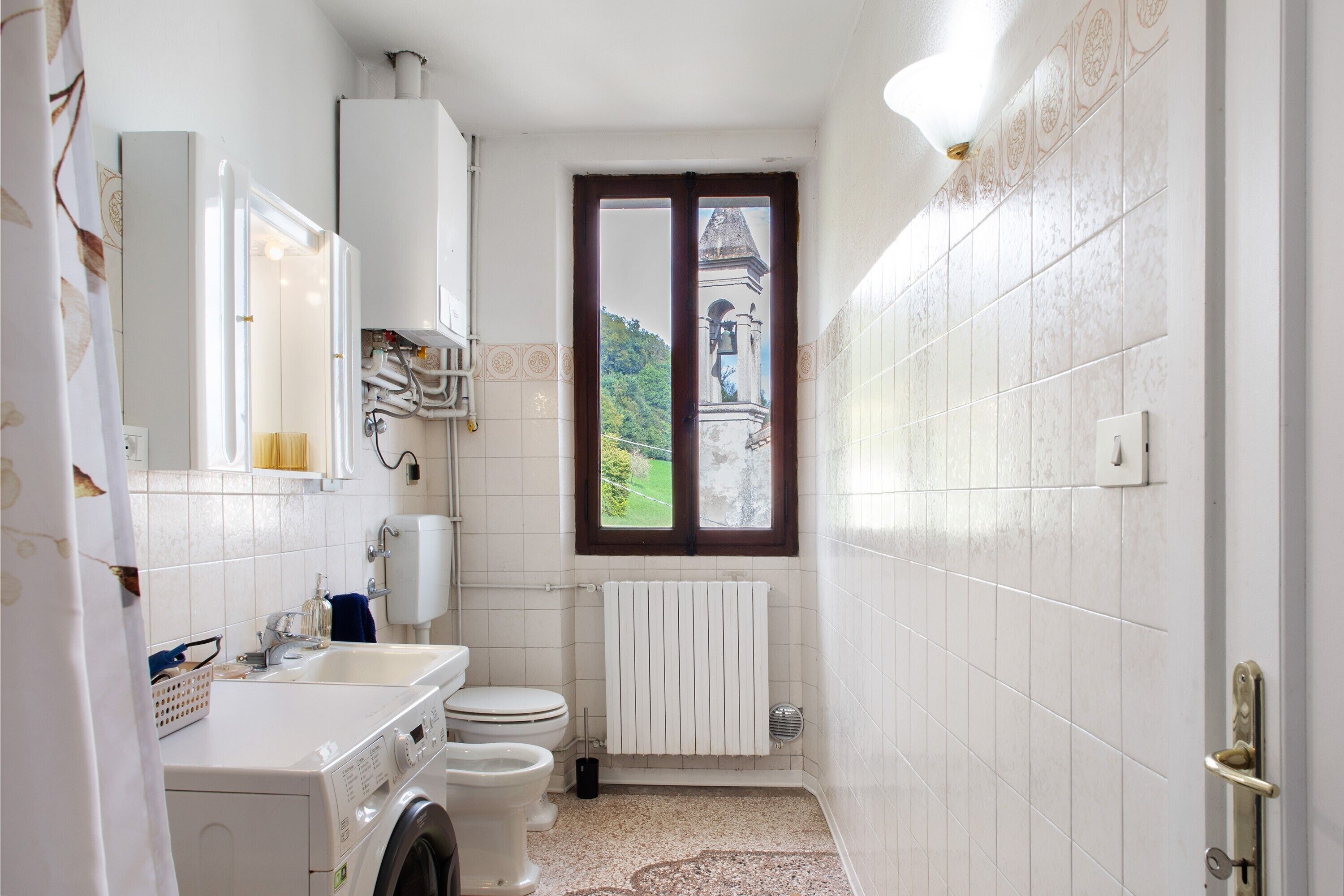 Ein kleines Badezimmer mit Waschmaschine, Toilette und einem Fenster mit Blick auf ein Gebäude.