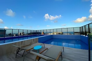 A rooftop pool with clear blue sky and clouds, surrounded by glass railings, and includes lounge chairs and a small table. - Compact Tambau  (João Pessoa)
