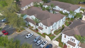Zona residencial amb diverses cases de dues plantes, cadascuna amb teulada marró i parets blanques. Hi ha diversos cotxes aparcats a la calçada, i la zona està envoltada de vegetació.