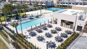 A rooftop pool area with multiple lounge chairs and tables, surrounded by a low fence and palm trees.