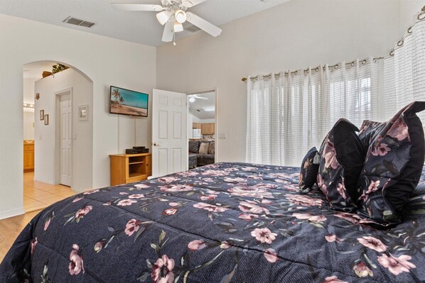 Un dormitorio con cubrecama estampado floral, ventilador de techo y vista a la cocina a través de una puerta abierta.