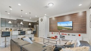 A modern living room with a sofa, coffee table, and a TV mounted on a wooden wall. There is a kitchen area with dark cabinets and a microwave.