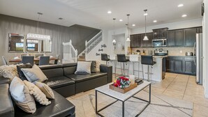 A modern living room with a black leather sofa, a coffee table, and a dining area with a white table and chairs. A kitchen with dark cabinets and stainless steel appliances is visible in the background.