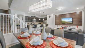 A modern dining area with a wooden table set for four, a kitchen in the background, and a living area with a TV.