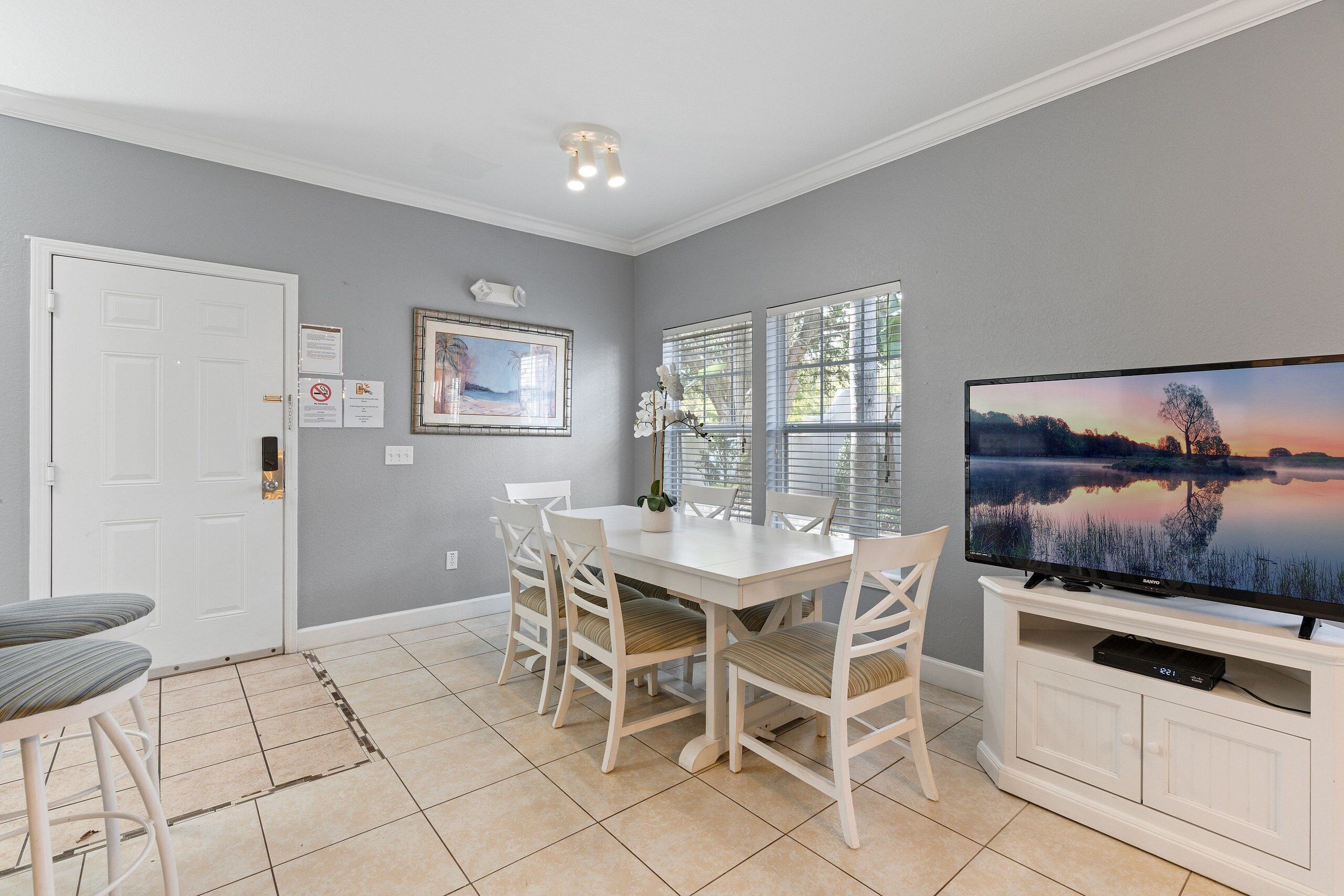 A dining area with a white table and chairs, a television on a stand, and a window with blinds.