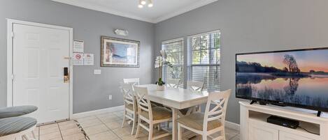 A dining area with a white table and chairs, a television on a stand, and a window with blinds.