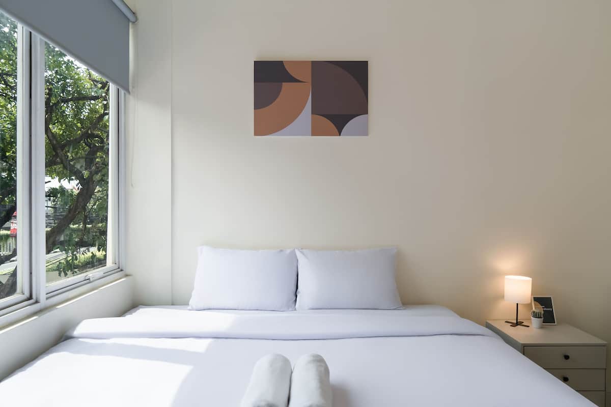 Ein Hotelzimmer mit einem Bett, einem Nachttisch mit Lampe und einem Fenster mit Blick auf Bäume.