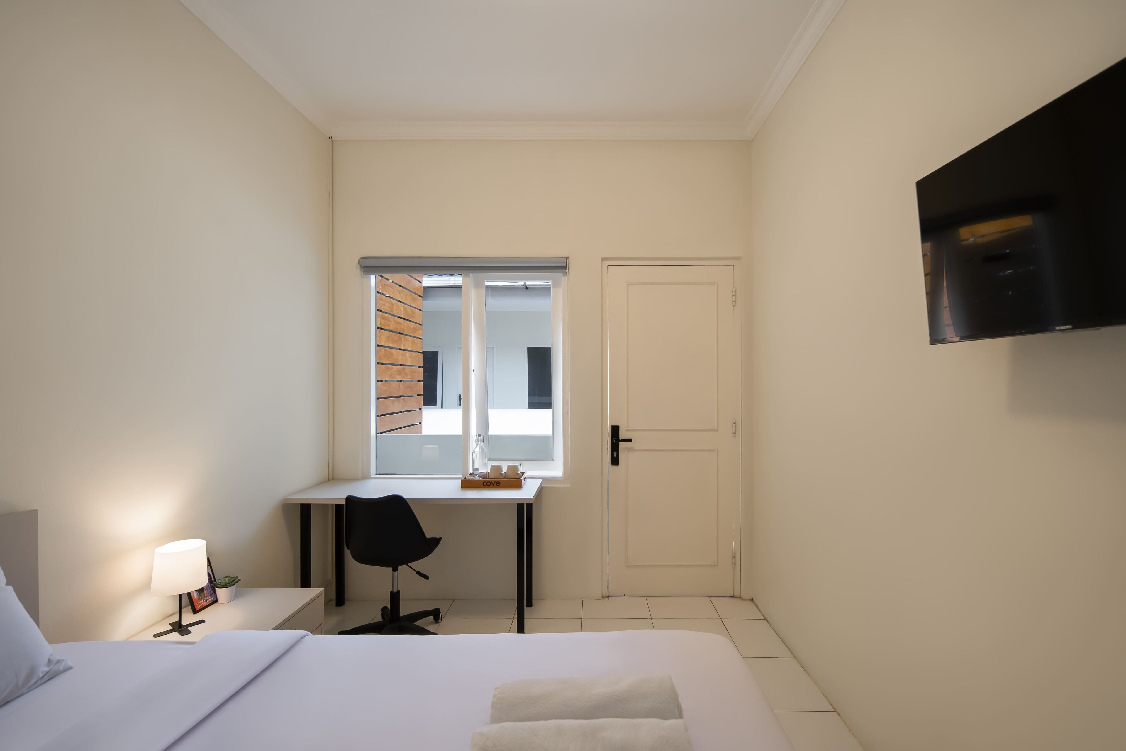 A hotel room with a bed, a desk, a chair, a TV mounted on the wall, and a window with a view of the outside.