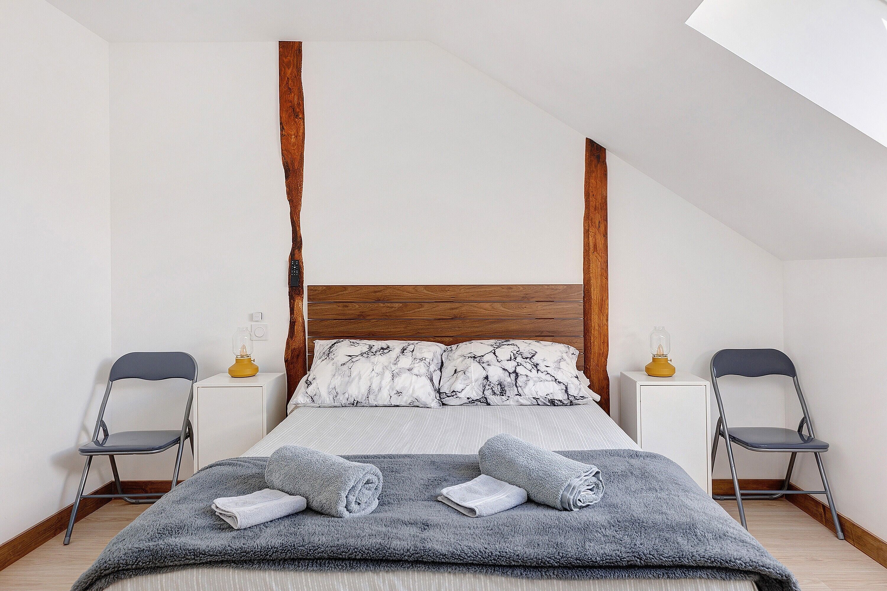 Ein Schlafzimmer mit einem hölzernen Kopfteil, einem Bett mit grauer Decke, zwei Nachttischen mit Lampen und zwei Klappstühlen.