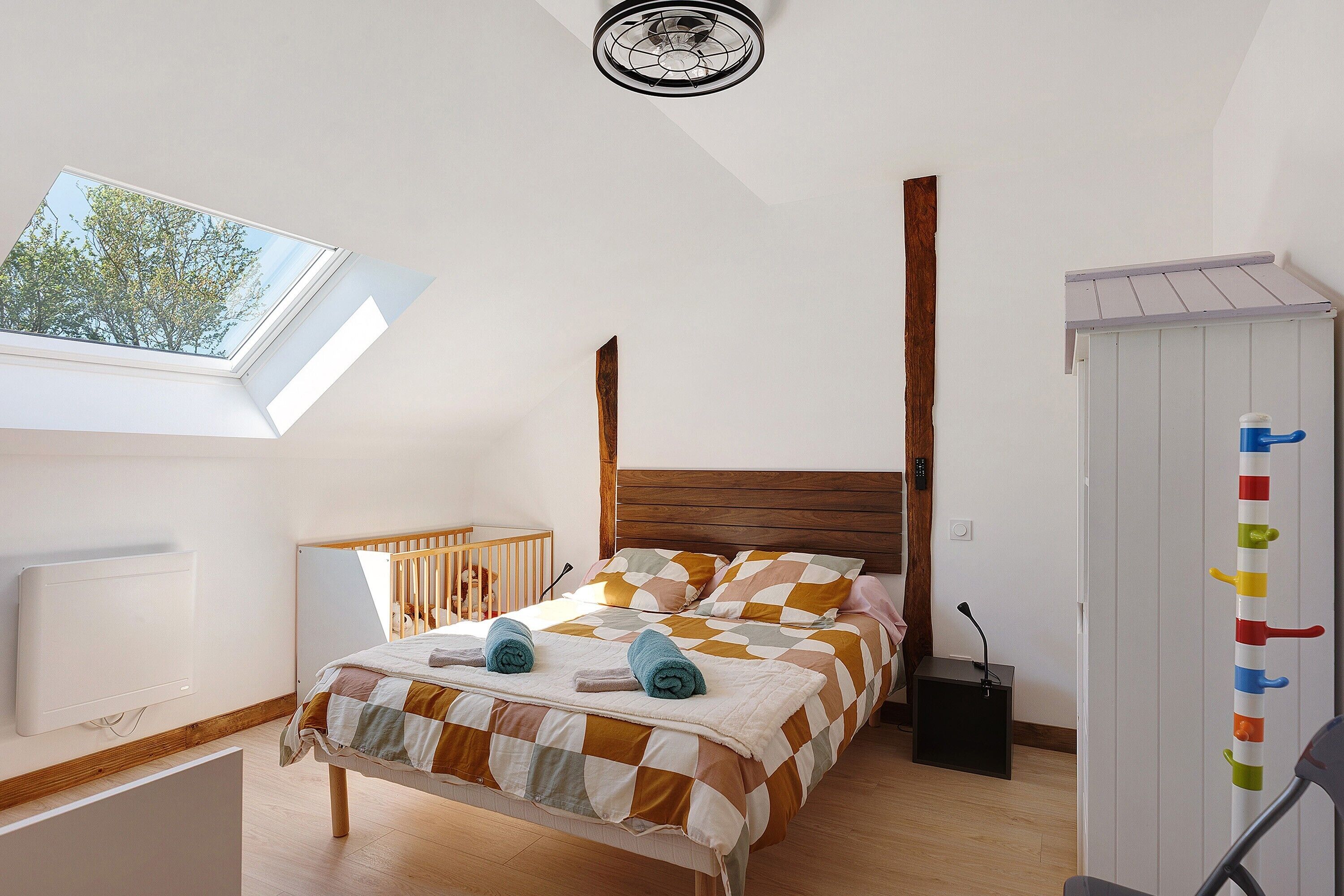 Une chambre avec un puits de lumière, une tête de lit en bois, un lit recouvert d’une couverture à carreaux et une table de chevet avec une lampe.