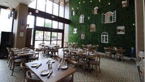 Ein Restaurant mit Holztischen und -stühlen, großen Fenstern und einer grünen Wand mit Fensterornamenten.