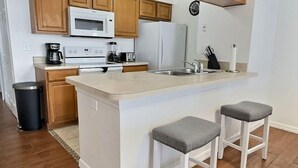 Una cocina con armarios de madera, una encimera blanca, microondas integrado, refrigerador y dos taburetes de bar.