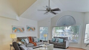 Un salón amplio con techo alto, ventilador de techo y un gran ventanal con persianas. Hay dos sofás y una mesa de centro.