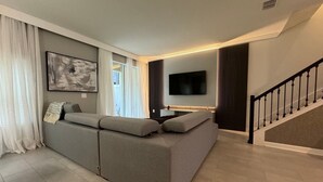A modern living room with a grey sofa, a flat-screen TV mounted on the wall, and a staircase with a black railing. - 5BR Storey Lake Home, Heated Pool (Kissimmee)