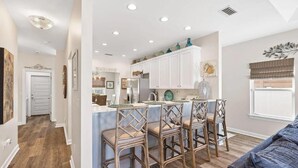 A modern kitchen with white cabinets, a central island with bar stools, and a dining area with a blue couch. - Palm Beach Oasis (Panama City Beach)