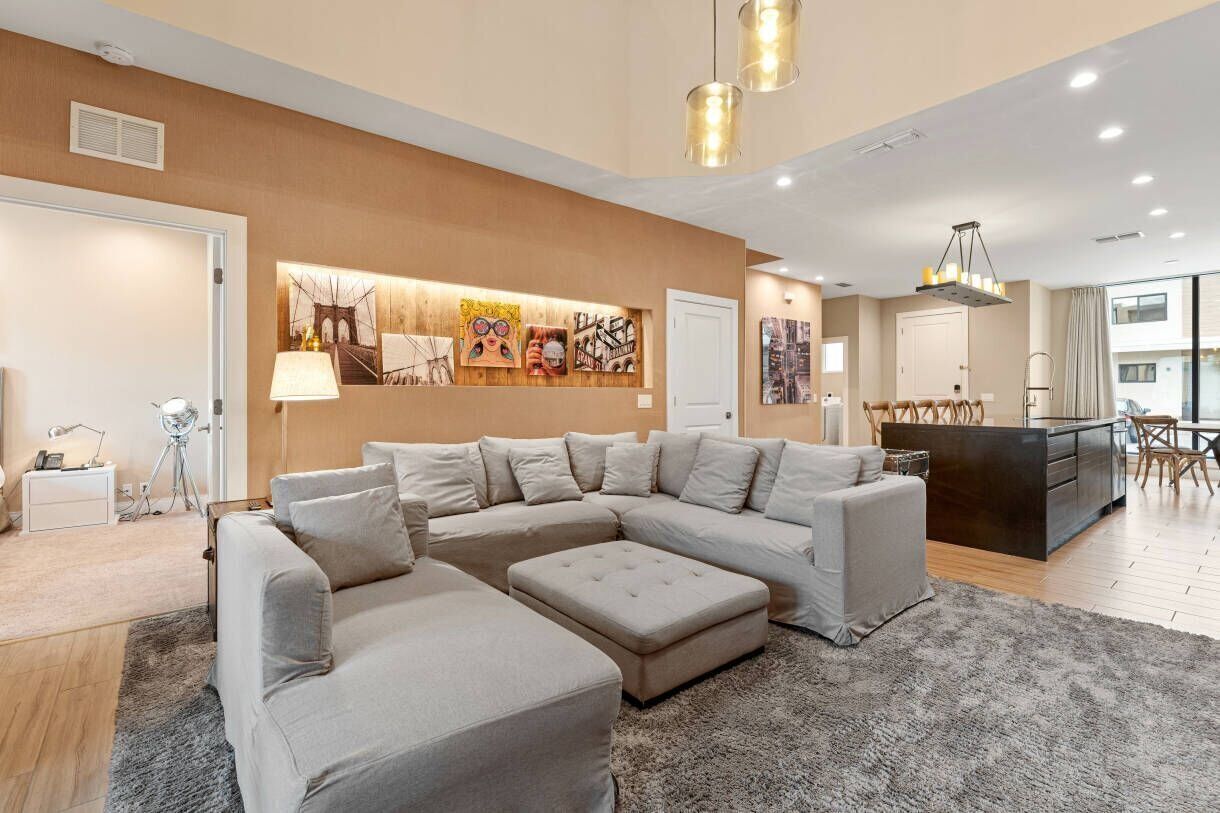 A modern living room with a sectional sofa, a small ottoman, and a coffee table. There is a dining area with a table and chairs, and a kitchen area with a black countertop and wooden cabinets. The room has a neutral color palette and features artwork on the walls.