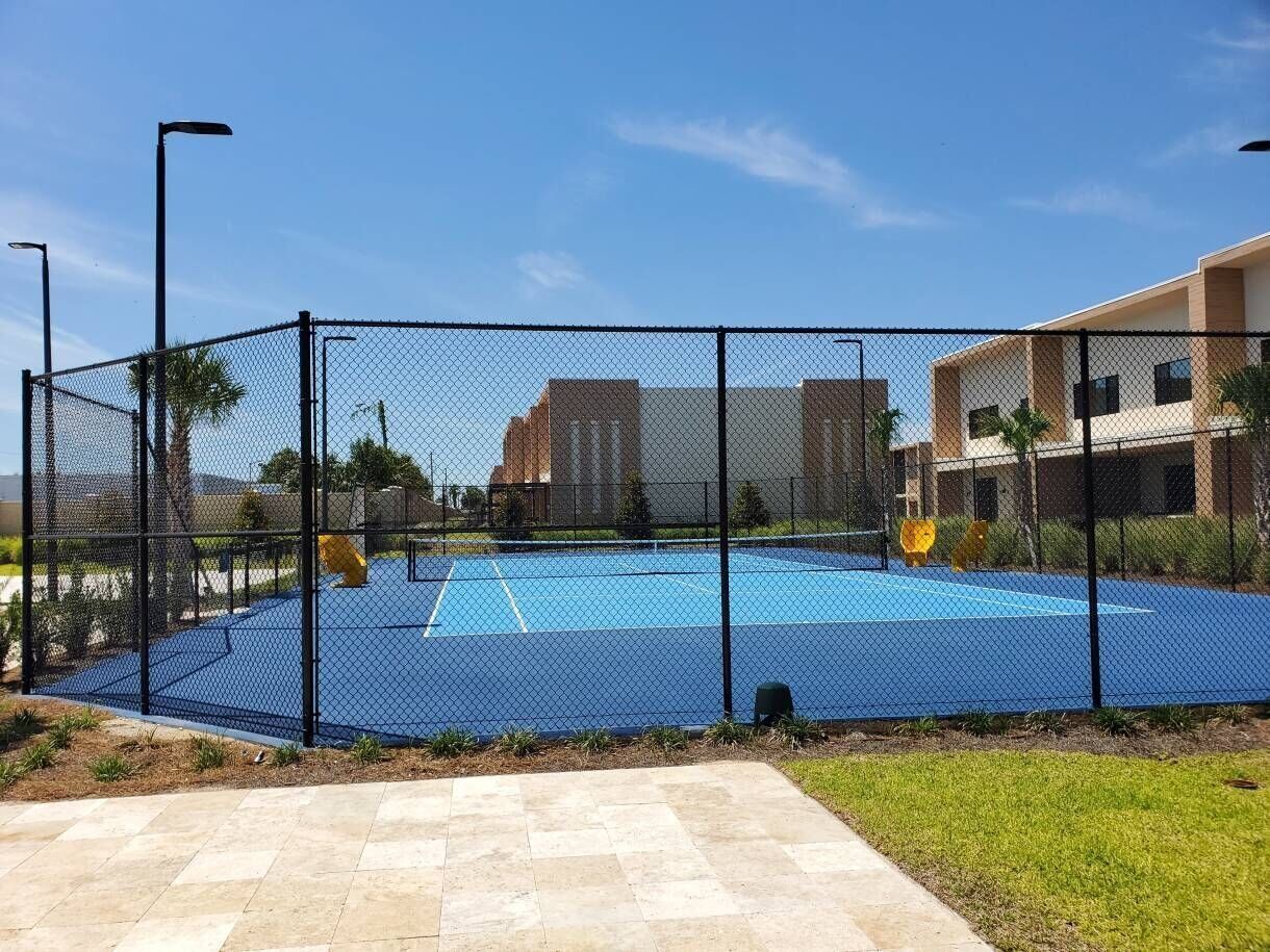 Una cancha de tenis al aire libre rodeada por una cerca de malla metálica, con un sendero pavimentado en el frente.