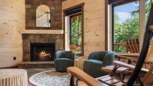 A cozy cabin interior with a stone fireplace, two blue armchairs, a wooden table, and a large window offering a view of trees. - NEW Luxury Cabin w/ Views*Private Pool*Game Room*Hot Tub (SEVIERVILLE)