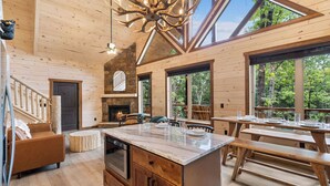 A spacious cabin interior with a large wooden dining table, a fireplace, and a view of the forest through large windows. - NEW Luxury Cabin w/ Views*Private Pool*Game Room*Hot Tub (SEVIERVILLE)