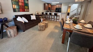 Un salon moderne avec un canapé marron foncé, une table basse et un coin repas comprenant une table en bois dressée pour quatre personnes.