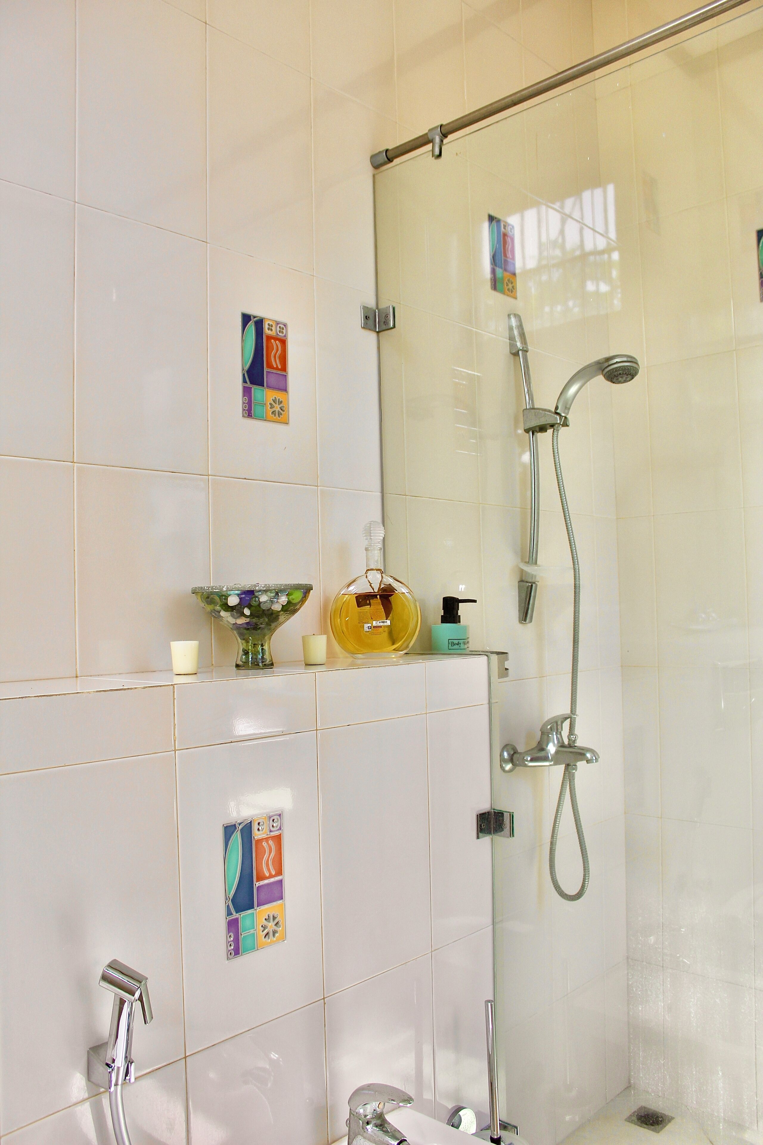A bathroom with a glass-enclosed shower, a handheld showerhead, and a wall-mounted faucet. There are decorative tiles on the wall and a small shelf with toiletries.