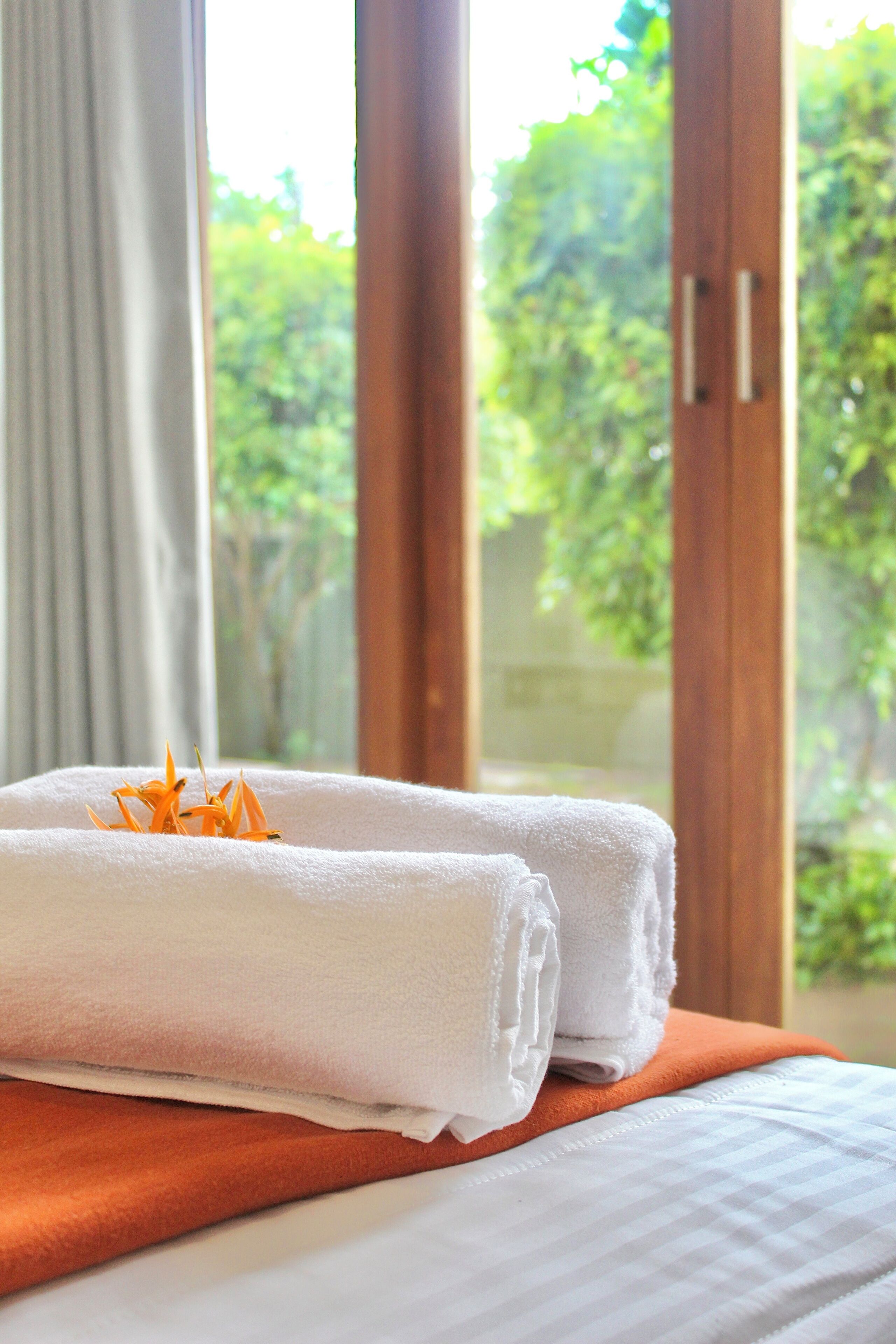 A neatly made bed with white and orange bedding, a window with curtains, and a view of greenery outside.