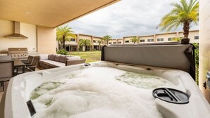 A hot tub with a view of a pool and palm trees, a barbecue area, and a seating area with a sofa and chairs.