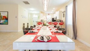 Een eetkamer met een lange tafel gedekt voor een maaltijd, een keuken op de achtergrond en een groot schilderij aan de muur.