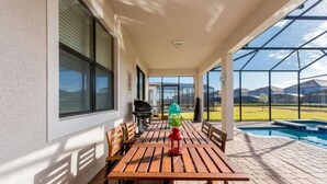 Een overdekt terras met een houten tafel en banken, een barbecue en een zwembad op de achtergrond.