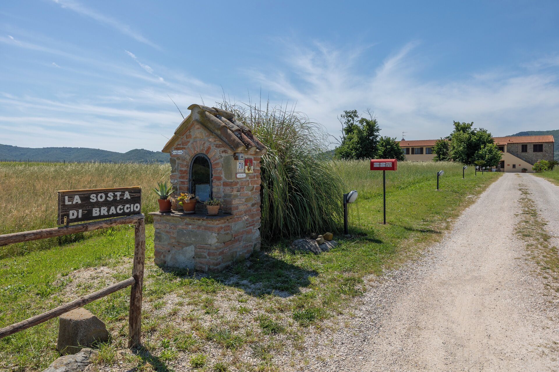 A rustic stone building with a small porch, a sign reading "LA SOSTA DI BRACCI", and a gravel road leading to a larger building in the background.
