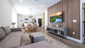 A modern living room with a grey sofa, a dining table with chairs, a TV mounted on a wooden wall, and a kitchen in the background.