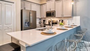 A modern kitchen with white cabinetry, stainless steel appliances, a central island with seating, and pendant lighting. - Stylish 2BR Condo | Avengers Room & Golf Course View (Kissimmee)