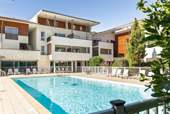 A swimming pool with lounge chairs, a multi-story building with balconies, and a clear sky.
